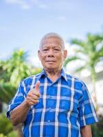 un mayor hombre usa un azul camisa y demostración dando pulgar arriba mientras en pie en un jardín. un mayor asiático hombre sano y tener positivo pensamiento. concepto de Envejecido personas y cuidado de la salud foto