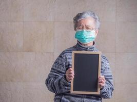 Portrait of an elderly Asian woman wearing a mask, holding a small blackboard and looking at the camera while standing with a light yellow background. Concept of aged people and healthcare photo