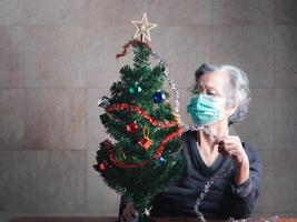 Elderly Asian woman wearing a mask and decorating the Christmas tree while sitting in a living room at home. Concept of aged people, healthcare and Christmas, and happy new year festival photo