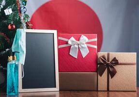 Small blackboard, mask, alcohol gel, and gift boxes on wooden table with a Christmas tree and Japan flag in the background. Space for text. Concept of Christmas and new year festival photo