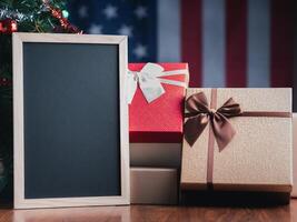 Small blackboard and gift boxes on wooden table with a Christmas tree and American flag in the background. Space for text. Concept of Christmas and new year festival photo