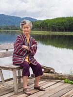 retrato de mayor asiático mujer brazos cruzado y mirando a el cámara mientras sentado en un de madera silla lado de el lago con montañas antecedentes. concepto de Envejecido personas y relajación foto