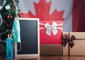 Small blackboard, mask, alcohol gel, and gift boxes on wooden table with a Christmas tree and Canada flag in the background. Space for text. Concept of Christmas and new year festival photo