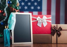 Small blackboard, mask, alcohol gel, and gift boxes on wooden table with a Christmas tree and American flag in the background. Space for text. Concept of Christmas and new year festival photo