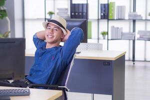 Young businessman wears a blue shirt and hat sitting on a chair in the office. He's smiling and happy to rest after work and holiday soon. Concept of business and relaxation photo