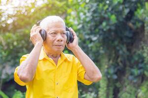 Senior man wearing wireless headphones listening to a favorite song and looking up while standing in a garden. Space for text. Concept of aged people and relaxation photo