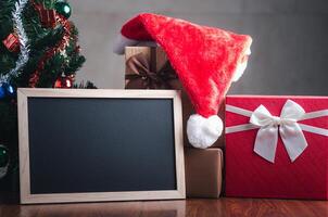 pequeño pizarra, un Papa Noel claus sombrero en regalo caja en de madera mesa con un Navidad árbol y borroso gris pared antecedentes. espacio para texto. concepto de Navidad y nuevo año festival foto