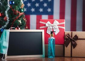 Small blackboard, mask, alcohol gel, and gift boxes on wooden table with a Christmas tree and American flag in the background. Space for text. Concept of Christmas and new year festival photo