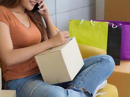 Asian businesswoman is an e-commerce entrepreneur using a smartphone and writes customer address on a parcel box while sitting on a sofa in the office. Concept of e-commerce photo