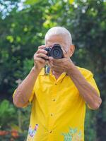 Senior man taking a photo by a digital camera in the park. An elderly Asian man wears a yellow shirt, happy when using a camera. Space for text. Concept of aged people and photography