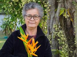 mayor asiático mujer vistiendo los anteojos, sonriente, participación flor amarillo, y mirando a el cámara mientras en pie en un jardín. concepto de Envejecido personas y cuidado de la salud foto