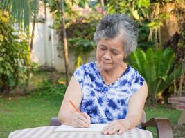 mayor mujer escritura un libro mientras sentado en un silla en el jardín. de cerca foto con Copiar espacio para texto. concepto de Envejecido personas y cuidado de la salud