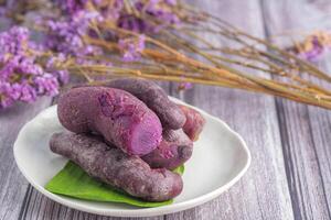 A pile of ripe purple yams is placed on a plate on a wooden table with blurred purple flowers in the background. Space for text. Concept of health fruit photo