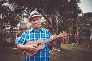 Portrait of an elderly Asian man wearing a hat playing the ukulele, smiling and looking at the camera while standing in a garden. Space for text. Concept of aged people and relaxation photo