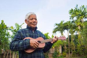 Portrait of an elderly Asian man playing the ukulele, smiling and looking away while standing in a garden. Space for text. Concept of aged people and relaxation photo