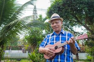 Portrait of an elderly Asian man wearing a hat playing the ukulele, smiling and looking at the camera while standing in a garden. Space for text. Concept of aged people and relaxation photo