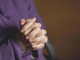 Hands of a senior woman praying while sitting on a chair in the living room. Close-up photo