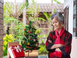 Portrait of senior woman arms crossed and looking at Christmas tree while standing near a Christmas tree at home photo