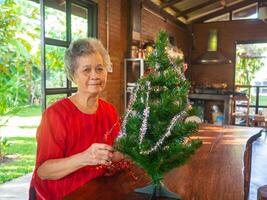 Senior Asian woman decorating the Christmas tree with white and red ribbons at home. Concept of aged people and Christmas and happy new year festival photo