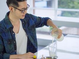 A young man wearing glasses pouring milk into a glass after breakfast while sitting on a chair in the kitchen room. Space for text. Selective focus. Health food and drink concept photo
