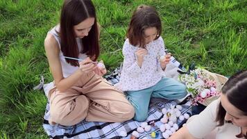 un familia de Tres muchachas de diferente siglos y europeo apariencia son sentado en el jardín en el verde césped y son comprometido en pintura Pascua de Resurrección huevos. Pascua de Resurrección tradiciones primavera día festivo. video