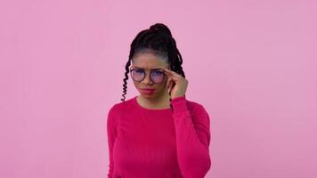 A cute African-American girl in pink clothes looks thoughtfully and seriously at the camera. Teen girl standing on a solid pink background video