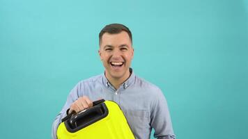 Happy positive man in a blue shirt happily shows off a stylish yellow plastic suitcase in his hands. Business man on a bright blue background video