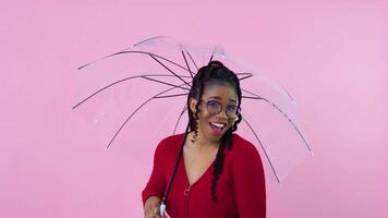 Nice young african american girl having fun with transparent umbrella in photo studio. Teen girl in red clothes holding an umbrella on a solid pink background video