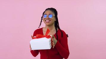 linda joven africano americano niña bailando con un regalo. hembra estudiante en rojo ropa sostiene un blanco caja con un rojo cinta. San Valentín día regalo video