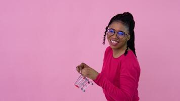 linda alegre joven africano americano niña en rosado ropa participación un juguete mini compras carro. adolescente niña ama de casa principiante en pie en un sólido rosado antecedentes video
