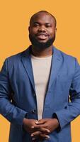 Vertical Portrait of joyous african american man holding hands together, smiling, looking pleased, studio background. Upbeat BIPOC person elegantly dressed exulting, feeling satisfied, camera B video