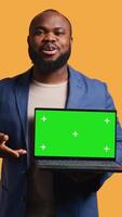 Vertical Smiling man holding isolated screen laptop, doing recommendation, talking with viewers. Cheerful BIPOC person pointing towards chroma key notebook, studio background, camera B video