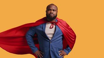 African american superhero flying with red cape, isolated over studio background, flexing muscles. Man wearing cloak posing as hero in costume showing courage and strength, camera B video