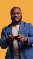 Vertical Joyous BIPOC gamer celebrating after winning gaming console game, studio background. Delighted man bragging after being victorious in game, defeating all enemies using gamepad, camera B video