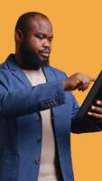 Vertical Man happily scrolling on tablet touchscreen to check social media feed. African american person browsing internet websites, entertained by content, studio background, camera B video