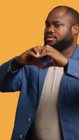 Vertical Portrait of jolly friendly african american man doing heart symbol shape gesture with hands. Cheerful nurturing BIPOC person showing love gesturing, isolated over studio background, camera A video