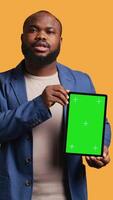 Vertical Portrait of african american man doing influencer marketing using green screen tablet, studio background. Smiling BIPOC person holding empty copy space mockup device, camera B video