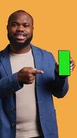 Vertical Smiling african american man presenting cellphone with green screen display, isolated over studio background. Cheerful BIPOC person creating promotion with blank copy space mockup phone, camera B video