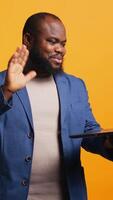 Vertical Cheerful employee greeting coworkers during teleconference meeting using laptop , studio background. Smiling worker waving hand, saluting colleagues during internet call, camera B video