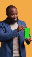 Vertical Cheerful african american man recommending green screen smartphone, laughing, studio background. Joyous BIPOC person presenting mockup mobile phone, giving positive feedback, camera B video