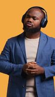 Vertical Relaxed african american man having fun listening to music on headphones during time off. Carefree BIPOC person enjoying songs and singing lyrics, isolated over studio background, camera B video