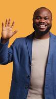 Vertikale afrikanisch amerikanisch Mann fröhlich tun Anrede Hand Geste. Porträt von glücklich bipoc Person erziehen Arm zu grüßen jemand, gestikulieren, isoliert Über Gelb Studio Hintergrund, Kamera ein video