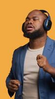 Vertical Happy african american person having fun, dancing on rhythm, listening to headphones. Cheerful BIPOC man entertaining himself doing funky movements on music, studio background, camera A video