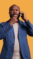 Vertical African american man having thoughtful conversation with friend during telephone call, studio background. Pensive BIPOC person talking on phone with mate, doing thinking gesturing, camera B video