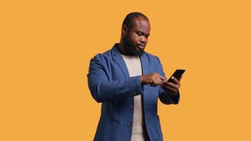 Portrait of joyous man holding cellphone celebrating, throwing fists in air and screaming. Delighted african american person reading good news on mobile phone, cheering, studio background, camera B video