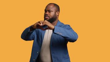 Portrait of jolly friendly african american man doing heart symbol shape gesture with hands. Cheerful nurturing BIPOC person showing love gesturing, isolated over studio background, camera A video