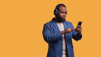 sonriente africano americano hombre escuchando a música en Bluetooth auriculares conectado a teléfono inteligente relajado bipoc persona seleccionando canciones desde lista de reproducción, aislado terminado estudio fondo, cámara si video