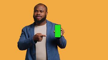 sorridente africano americano homem apresentando celular com verde tela mostrar, isolado sobre estúdio fundo. alegre bipoc pessoa criando promoção com em branco cópia de espaço brincar telefone, Câmera uma video