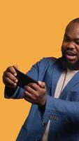 Vertical African american man playing games on cellphone in landscape mode, enjoying leisure time. Gamer enjoying game on mobile phone, having fun, studio background, camera A video