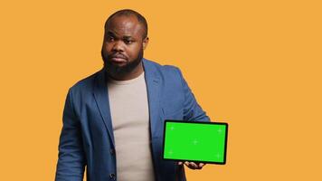 Upset african american man shows thumbs down sign gesturing holding isolated screen tablet, disapproving. BIPOC person does rejection hand gesture regarding mockup device, studio background, camera A video